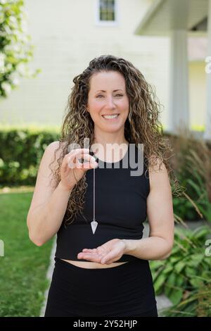 Giovane donna maestra di Reiki sorridente mentre tiene in mano il pendolo di cristallo Foto Stock