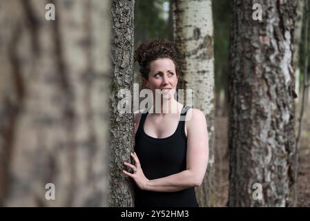 Giovane donna con espressione gioiosa si appoggia contro l'albero nella foresta Foto Stock