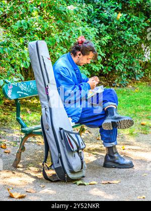 Giovane musicista con strumento nel caso riposi e rotolare una sigaretta nel giardino di Francesco Sicard - Tours, Indre-et-Loire (37), Francia. Foto Stock