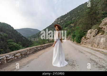 Una donna con un vestito bianco si erge su una strada con montagne sullo sfondo Foto Stock