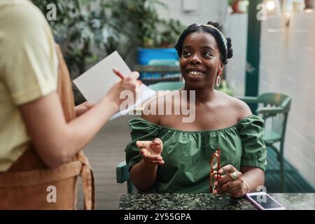 Donna afro-americana seduta a tavola e sorridente mentre fa l'ordine nel bar, cameriera irriconoscibile in piedi accanto al tavolo e prende appunti Foto Stock