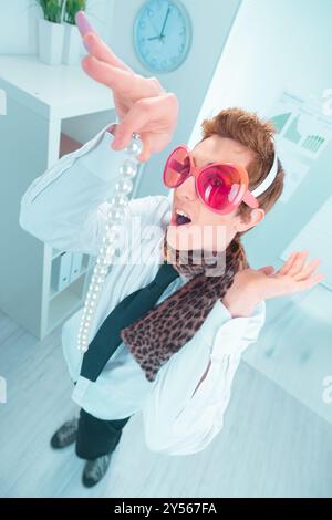 Un uomo d'affari in un buffo travestimento tiene una collana di perle, che sembra sorpreso e divertito in un ambiente d'ufficio Foto Stock