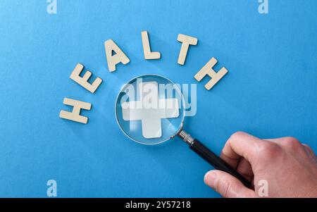 Informazioni mediche ed esame sfondo con mano che ingrandisce una croce medica con una lente d'ingrandimento e lettere con messaggio di salute su blu è Foto Stock