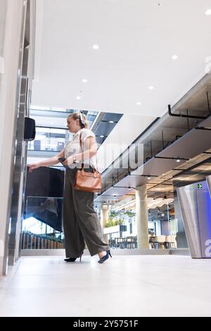 Premendo il pulsante dell'ascensore, una donna d'affari in ufficio che tiene in mano un tablet e una borsa Foto Stock