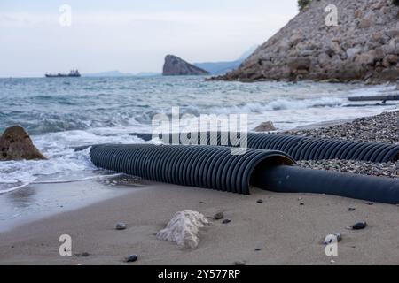 Tubi di drenaggio neri lavati a terra su una spiaggia rocciosa con onde dolci durante le ore diurne Foto Stock