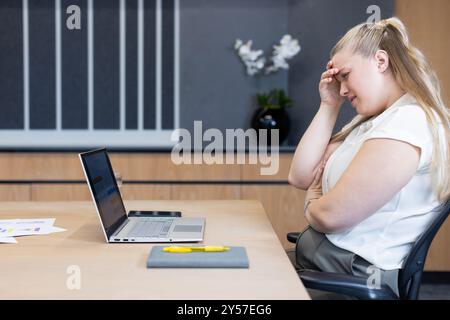Donna d'affari stressata seduta alla scrivania con laptop e documenti in ufficio Foto Stock