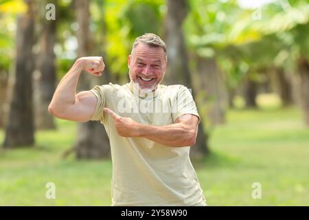 Un uomo allegro e maturo che si flette il braccio e punta al bicipite sorridendo, godendosi un momento di divertimento all'aperto in un ambiente verde e soleggiato. Foto Stock