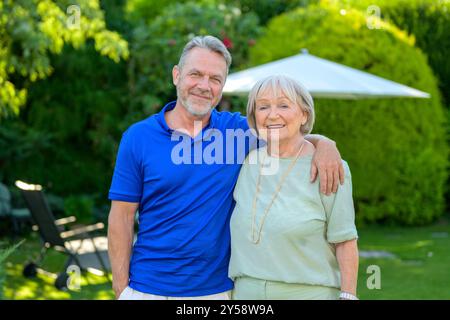 Un uomo maturo e una donna anziana in piedi vicini, sorridendo calorosamente e godendosi una giornata di sole in un lussureggiante giardino verde. Foto Stock