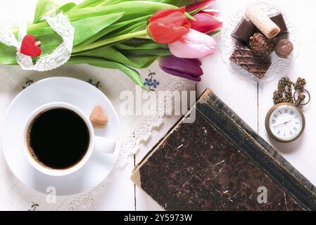 Romantico tavolo per la colazione con una tazza di caffè, dolci, un vecchio libro, un orologio tascabile vintage e un bouquet di tulipani legati a pizzi, su un tavolo bianco Foto Stock