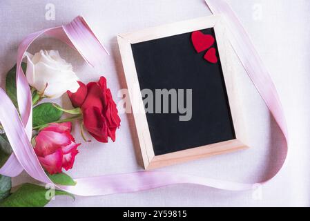 Immagine a tema di San Valentino con splendide rose legate con nastro rosa e una lavagna vuota decorata con due cuori rossi Foto Stock