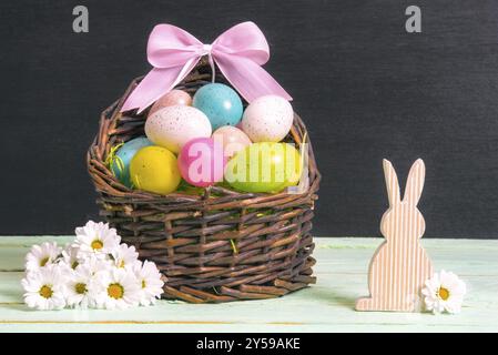 Cestino di vimini pieno di uova di Pasqua multicolore e fiocco rosa, circondato da margherite bianche e un coniglietto di legno, su un tavolo e una parete nera Foto Stock