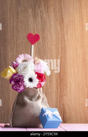 Tessera di San Valentino con un bellissimo bouquet di fiori in una borsa di iuta, con un cuore rosso e una scatola regalo blu, su un tavolo rosa e uno sfondo di legno Foto Stock