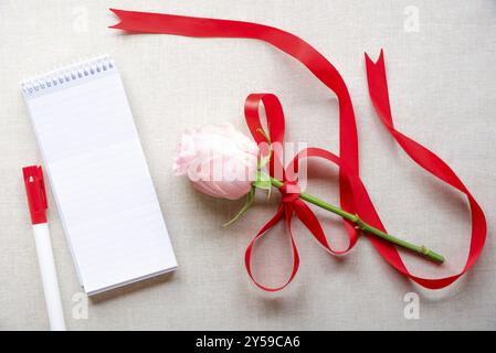 Splendida rosa legata a un nastro in raso rosso con fiocco, un taccuino a spirale non scritto a righe e un pennarello, su uno sfondo in tessuto vintage Foto Stock