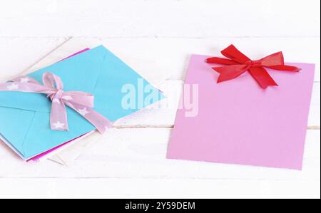 Mazzo di lettere multicolore legate insieme a un nastro rosa e inchino vicino a una nota di carta rosa vuota con un fiocco rosso, su uno sfondo di legno bianco Foto Stock
