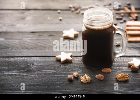 Grande Vaso in vetro pieno con caffè caldo e circondato da cannella a forma di stella i cookie, le noci e le mandorle in un rustico di tavoli in legno nero, sotto il mattino Foto Stock