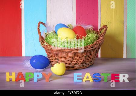 Cesto decorativo in vimini con uova dipinte e le parole Happy easter scritte con lettere in legno multicolore e una recinzione colorata sullo sfondo Foto Stock