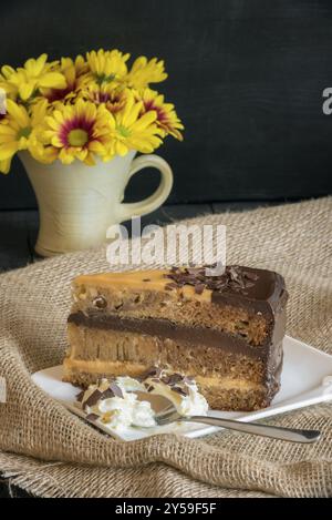 Fetta di torta brasiliana con ripieno brigadeiro al latte e al cioccolato, servita con panna montata, su un piatto bianco, tessuto di iuta e fiori gialli Foto Stock