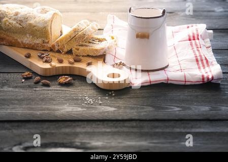 Gustosa torta fatta in casa con semi di papavero e noci, tagliata su una scavatrincee, vicino a un secchio di latte, su un tavolo di legno nero, alla luce del mattino Foto Stock