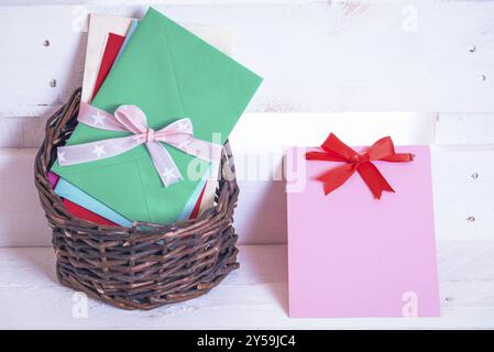 Pile di buste multicolore legate con nastro, esposte in un cestello di vimini e in una nota di carta con un fiocco rosso vicino, su sfondo bianco di legno Foto Stock