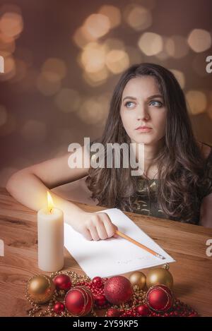 Bella giovane donna, con gli occhi blu, che pensa a cosa scrivere, a lume di candela, circondata da globi rossi e gialli e luci di Natale Foto Stock