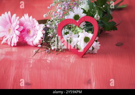 Immagine a tema d'amore con un cuore di legno rosso appoggiato su un mazzo di fiori, su un tavolo di legno rosso. Un concetto di relazione, amore, san valentino Foto Stock