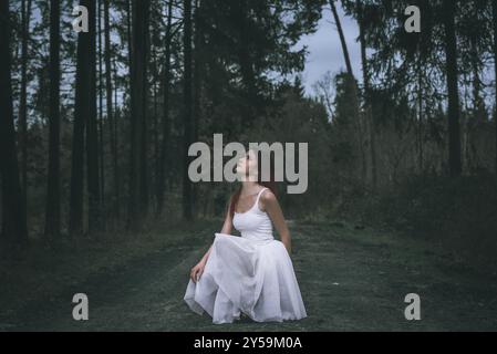 Bella donna con un'acconciatura ombre, vestita con una gonna in tutù bianca, in posizione squat, su una strada forestale, guardando in alto, in un bosco primaverile Foto Stock