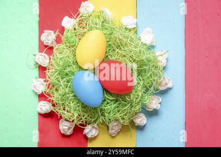 Allestimento pasquale con uova dipinte su erba decorativa, circondate da piccole rose bianche, esposte su uno sfondo da tavole di legno multicolore Foto Stock