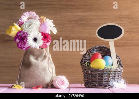 Bouquet di fiori in un sacco di iuta e un cesto di vimini con uova dipinte e uno striscione di legno bianco, su un tavolo rosa, con una parete di legno Foto Stock