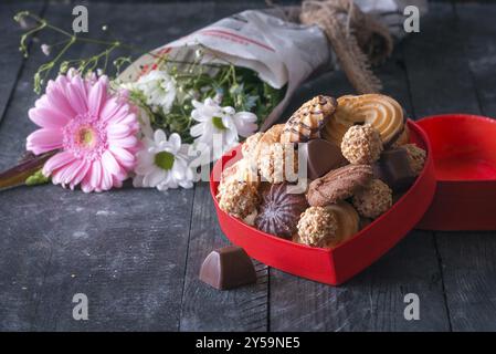 Regali foto a tema con un riquadro rosso pieno di biscotti, cioccolatini e un mazzo di fiori avvolti in un giornale in background su un vintage ta in legno Foto Stock