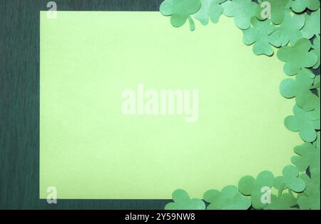 Design del biglietto d'auguri di San Patrizio con molti intagli di carta intorno a un foglio di carta giallo vuoto, visualizzato su uno sfondo verde scuro in legno Foto Stock