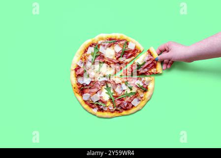 Vista dall'alto con una pizza al prosciutto fatta in casa isolata su uno sfondo verde. La mano di una donna che prende una fetta di pizza Foto Stock