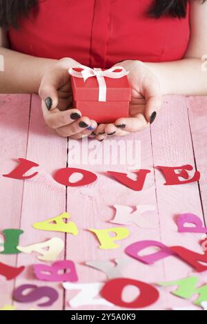 Le mani di una donna tengono una scatola regalo rossa su un tavolo di legno rosa, pieno di lettere di carta multicolore e la parola amore scritta da loro Foto Stock