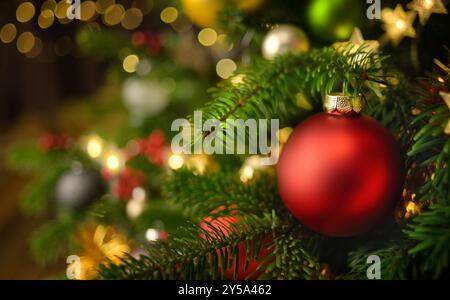 Primo piano di un pallone rosso di Natale appeso su un abete decorato, con molti ornamenti e luci lucenti che creano il bokeh Foto Stock