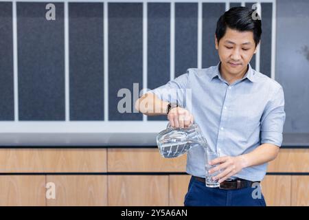 Versare l'acqua dalla caraffa al vetro, uomo asiatico in abbigliamento da lavoro Foto Stock