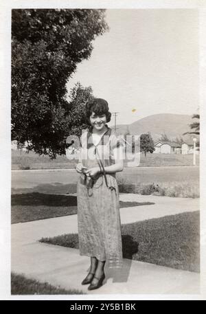 Una giovane donna sorride per la macchina fotografica mentre sta in piedi su un marciapiede soleggiato, vestito con un abito leggero e fantasia. La foto cattura un momento di gioia in un tranquillo quartiere periferico, probabilmente degli anni '1920 o '1930 nel sud della California. Foto Stock