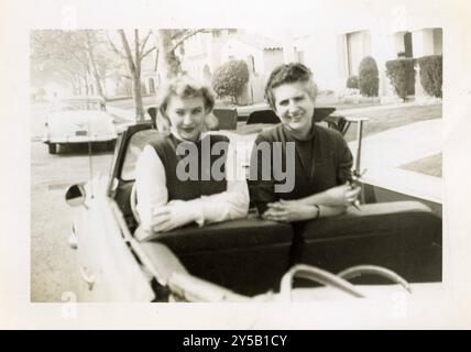 Due donne che si divertono a guidare su una cabriolet, probabilmente degli anni '1940 o '1950, parcheggiate in una tranquilla strada di periferia costeggiata da auto d'epoca. Foto Stock