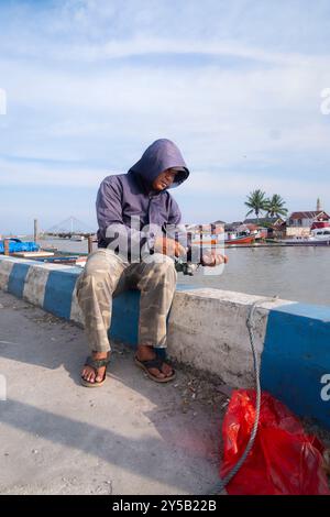 Balikpapan, Borneo orientale, Indonesia - 19 settembre. Un uomo siede su un muro vicino all'acqua, indossando una giacca con cappuccio e infradito Foto Stock