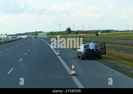 Gliwice, Polonia - 15 giugno 2023: Un uomo vestito con un abbigliamento formale si trova accanto alla sua auto con la porta aperta, guardando verso il motore su un alto tranquillo Foto Stock