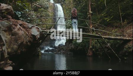Una giovane donna si gode la vista di una cascata in una foresta pluviale tropicale mentre si trova su un ponte di bambù. E' circondata da una vegetazione lussureggiante e dal suono dell'acqua a cascata Foto Stock