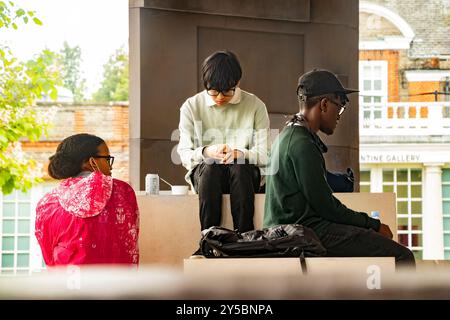 Tre giovani siedono in un ambiente all'aperto, ognuno impegnato nella propria attività, con un edificio storico sullo sfondo che sembra annoiato e depresso Foto Stock