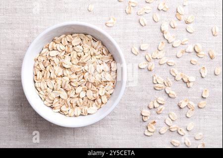 Fiocchi d'avena arrotolati, fiocchi d'avena arrotolati in una ciotola bianca su lino. Le farine d'avena cotte e cotte a vapore sono state arrotolate in fiocchi e leggermente tostate. Cibo integrale. Foto Stock