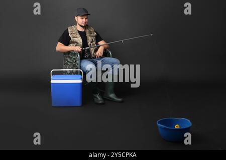 Pescatore con canna da pesca e scatola frigo che pesca dal ciotola sulla sedia su sfondo nero Foto Stock