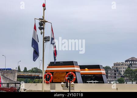 Bandiere olandesi sull'asta della nave contro il cielo grigio, tetto del flybridge, due salvagente arancioni, parte di un ponte di veicoli ed edifici sullo sfondo, giornata nuvolosa Foto Stock