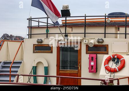 Flybridge sul ponte della nave, porta di legno, scala, bandiera olandese, tubo e salvagente arancione appeso al muro, giornata nuvolosa a Maastricht, Limburgo meridionale, Paesi Bassi Foto Stock