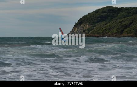 Uomo con windsurf affronta le onde al tramonto nel Golfo di Baratti, Piombino, Livorno, Italia Foto Stock