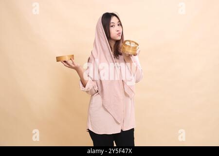 La musulmana asiatica indonesia bacia il dimsum guarda la macchina fotografica e la sua mano porta una ciotola di legno sul fondo contenente il dimsum. Foto Stock