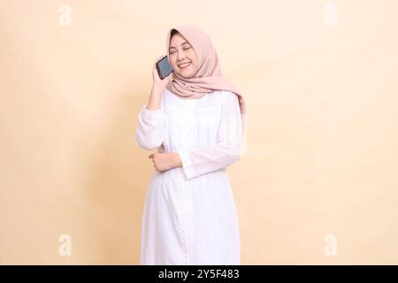 Donna musulmana asiatica che indossa un hijab, candida, sorridente, ridendo, tenendo in mano un gadget cellulare mentre chiama qualcuno. Stile di vita, tecnologia e. Foto Stock