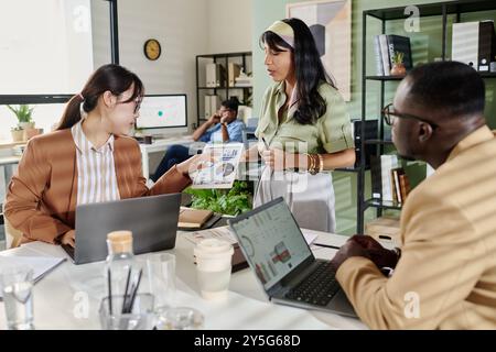 Gruppo di professionisti diversi che lavorano al progetto in un ufficio moderno, discutendo documenti con computer portatili aperti sulla scrivania che mostrano dati e grafici Foto Stock