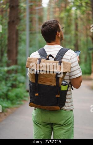 L'uomo che indossa uno zaino e tiene in mano uno smartphone cammina lungo un sentiero nella foresta, navigando attraverso la natura in un giorno d'estate. Foto Stock