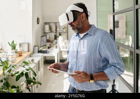 Uomo barbuto che utilizza un visore per la realtà virtuale mentre tiene il tablet in un ufficio arioso con piante e grandi finestre, profondamente impegnato con contenuti virtuali interattivi Foto Stock